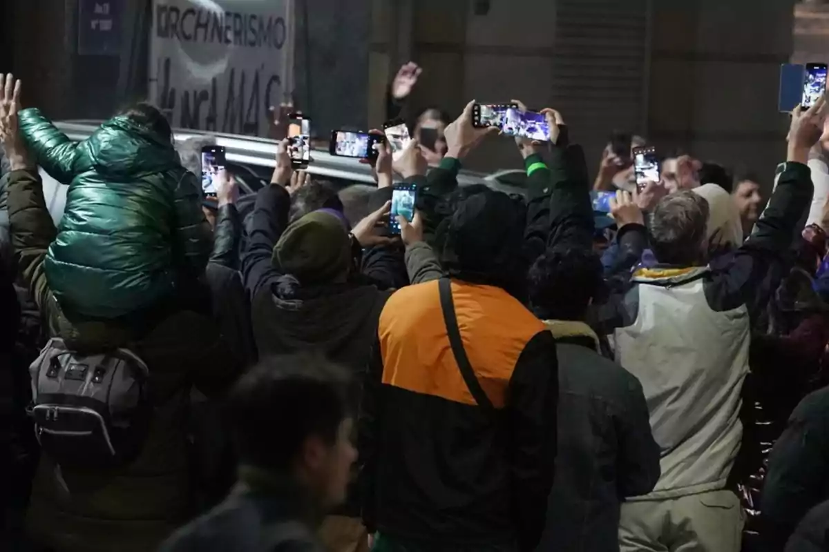 A group of people with their backs turned hold up their cell phones to take photos or record videos at a nighttime event, while one person carries a child on their shoulders.
