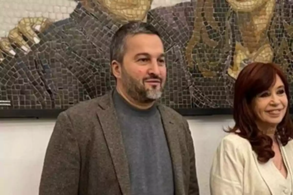 Two people pose in front of a mosaic mural; both are smiling and wearing formal clothing. Two people pose in front of a mosaic mural; both are smiling and wearing formal clothing.