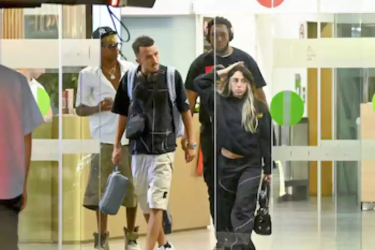 A group of people is leaving a building with glass doors; a woman in front is wearing dark clothes and carrying a bag, while three men follow her, one of them with headphones and another with a cap and sunglasses.