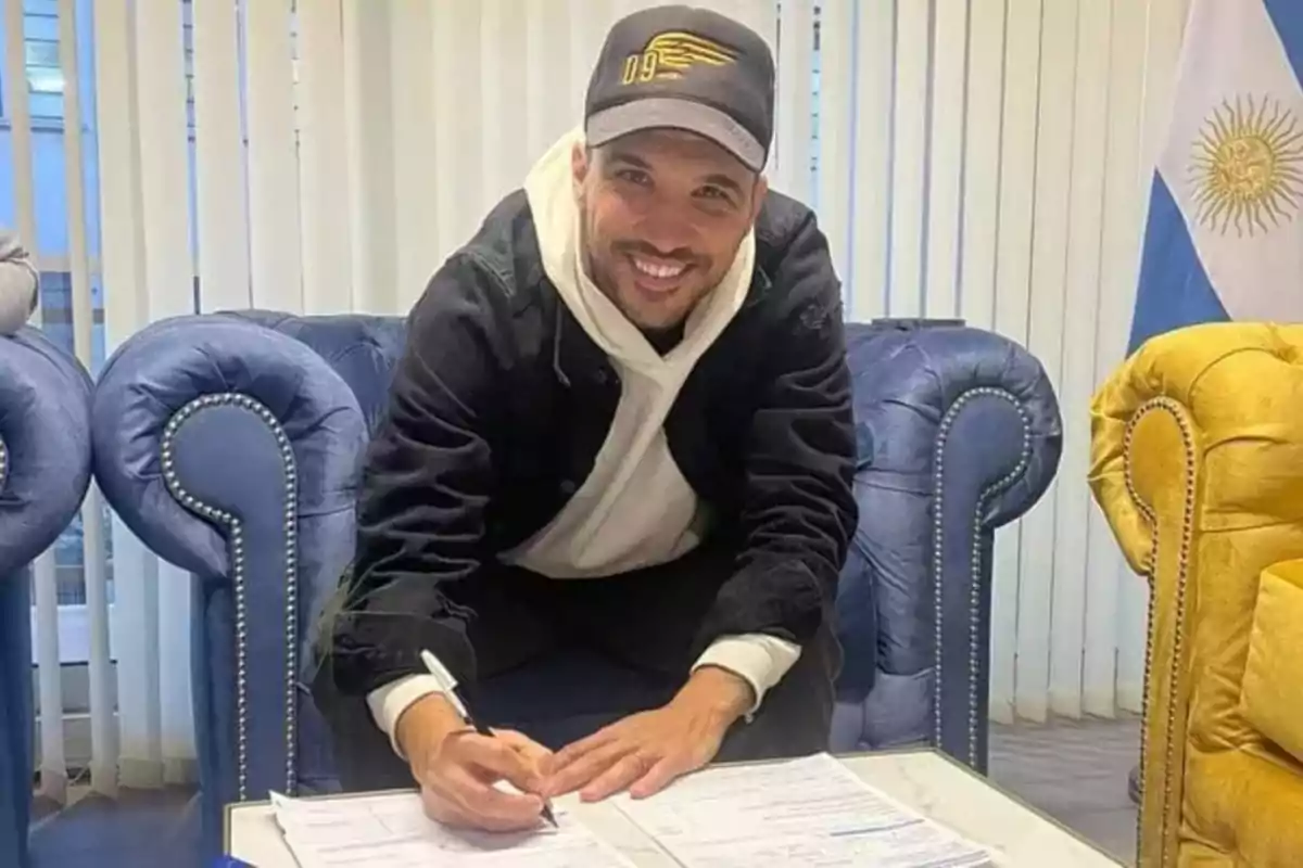 Hombre sonriente con gorra y sudadera firmando documentos en una mesa entre sillones azules y una bandera de Argentina al fondo Hombre sonriente con gorra y sudadera firmando documentos en una mesa entre sillones azules y una bandera de Argentina al fondo