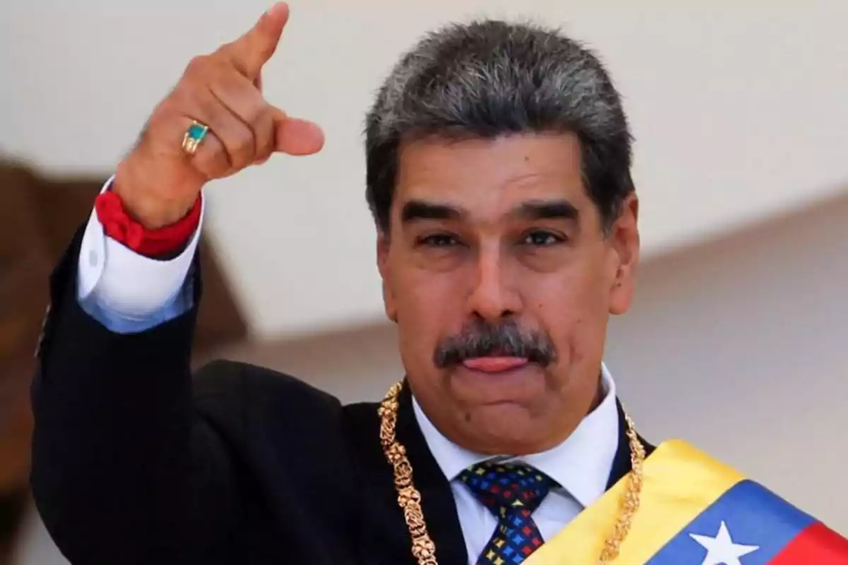 Hombre de cabello canoso y bigote usando banda presidencial y traje oscuro mientras señala con la mano y saca la lengua