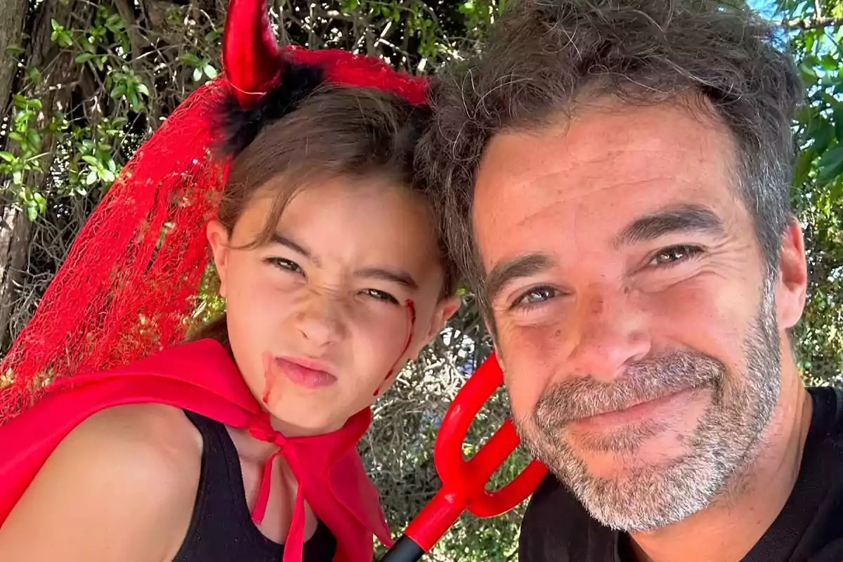 Niña disfrazada de diablo con capa y cuernos rojos junto a un hombre sonriendo, ambos al aire libre
