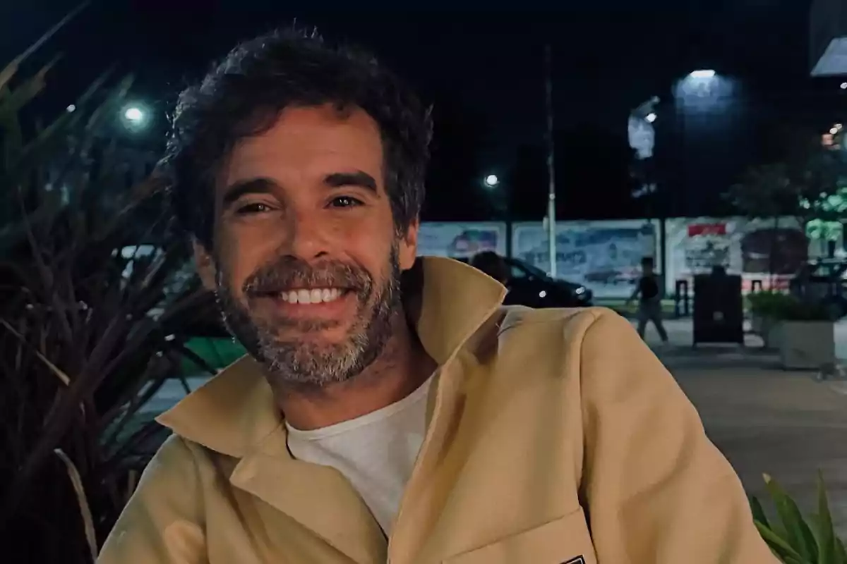 Hombre de cabello rizado y barba sonríe mientras está sentado al aire libre en una noche iluminada por luces urbanas