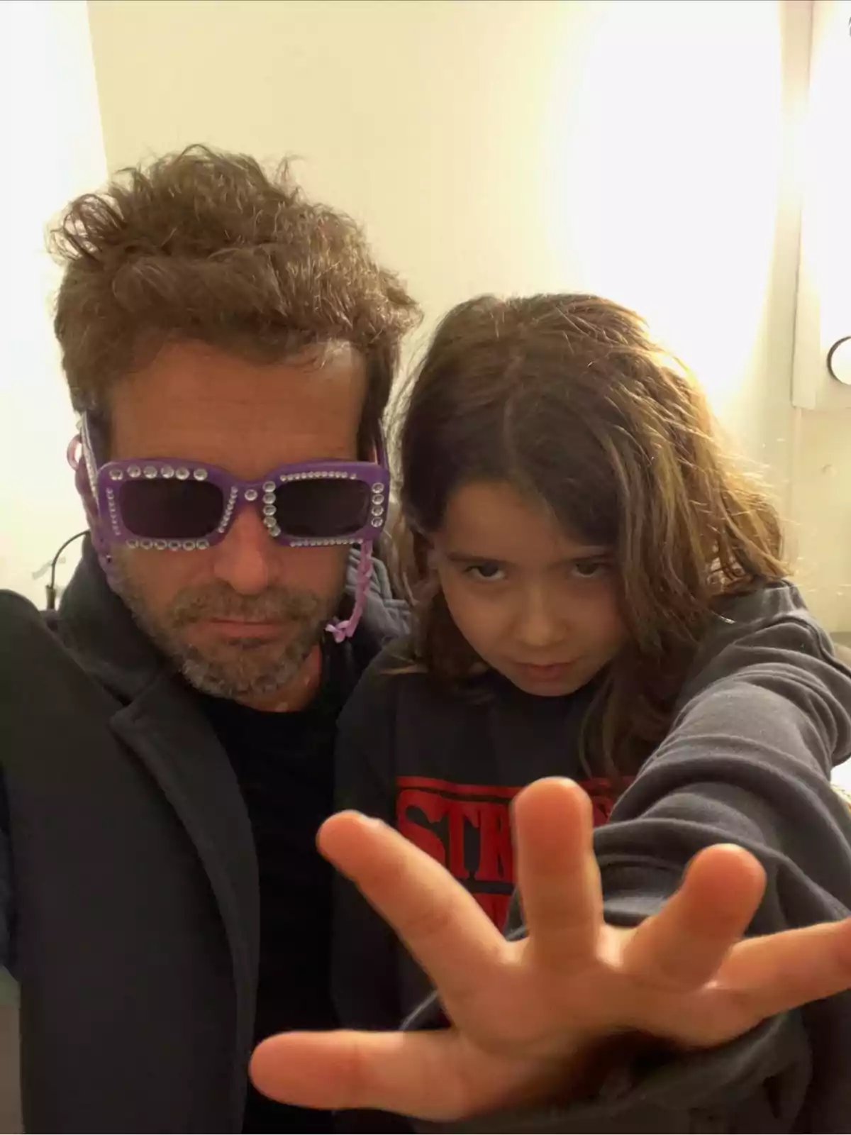 Un hombre con gafas moradas decoradas y una niña con expresión seria extiende la mano hacia la cámara, ambos posando en un ambiente interior iluminado. Un hombre con gafas moradas decoradas y una niña con expresión seria extiende la mano hacia la cámara, ambos posando en un ambiente interior iluminado.