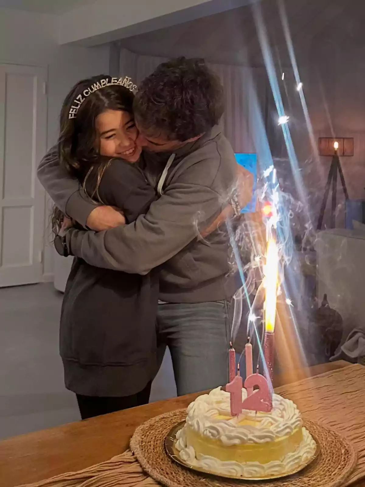 Una niña sonriente con una diadema de feliz cumpleaños recibe un abrazo cariñoso de un hombre mientras una tarta con velas encendidas y el número 12 está sobre la mesa Una niña sonriente con una diadema de feliz cumpleaños recibe un abrazo cariñoso de un hombre mientras una tarta con velas encendidas y el número 12 está sobre la mesa