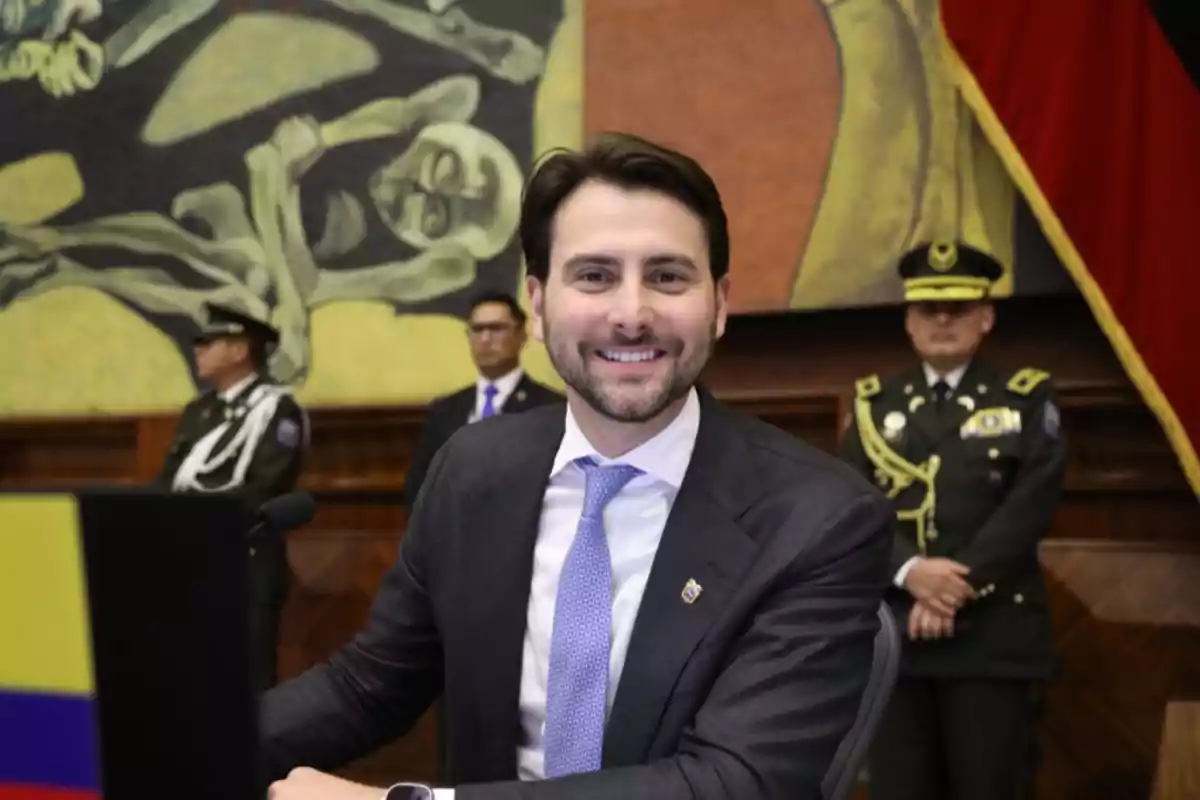 Hombre joven con traje y corbata sonríe frente a una mesa, al fondo hay dos oficiales uniformados y un mural artístico