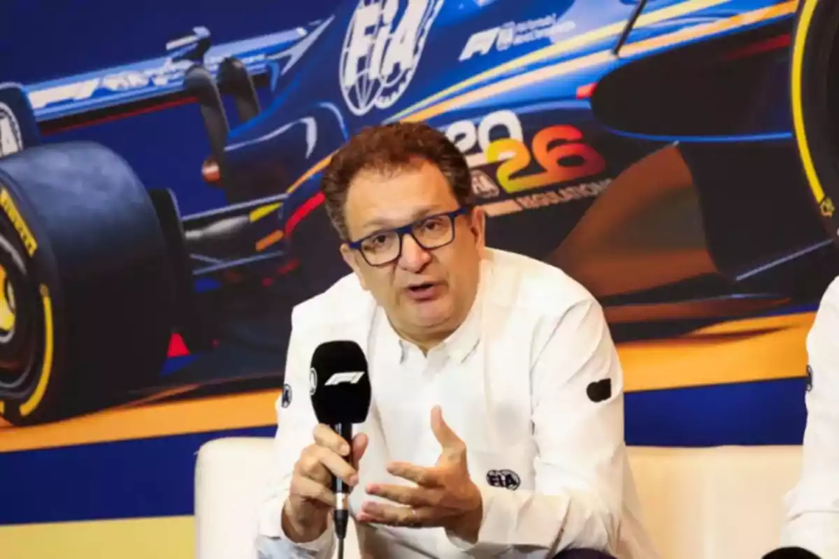 Man with glasses and a white jersey speaking at a Formula 1 press conference with a microphone in his hand and a race car in the background Man with glasses and a white jersey speaking at a Formula 1 press conference with a microphone in his hand and a race car in the background