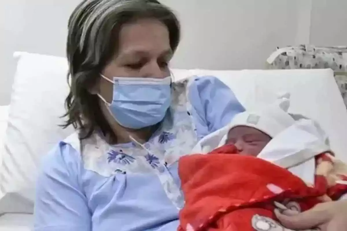 Woman wearing a face mask holding a newborn wrapped in a red blanket on a hospital bed Woman wearing a face mask holding a newborn wrapped in a red blanket on a hospital bed