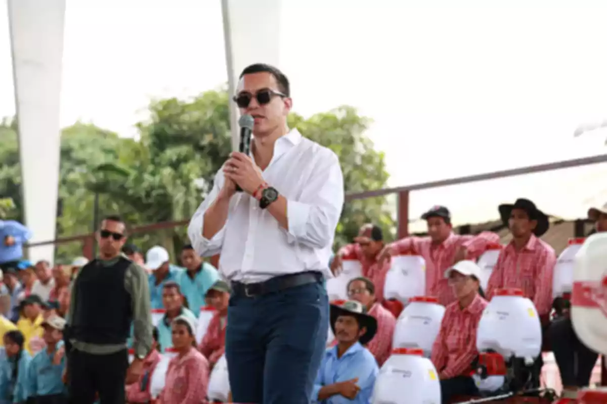 Hombre con camisa blanca y gafas de sol hablando por micrófono frente a un grupo de personas sentadas que llevan camisas a cuadros y sombreros, con mochilas fumigadoras blancas a su lado en un evento al aire libre.