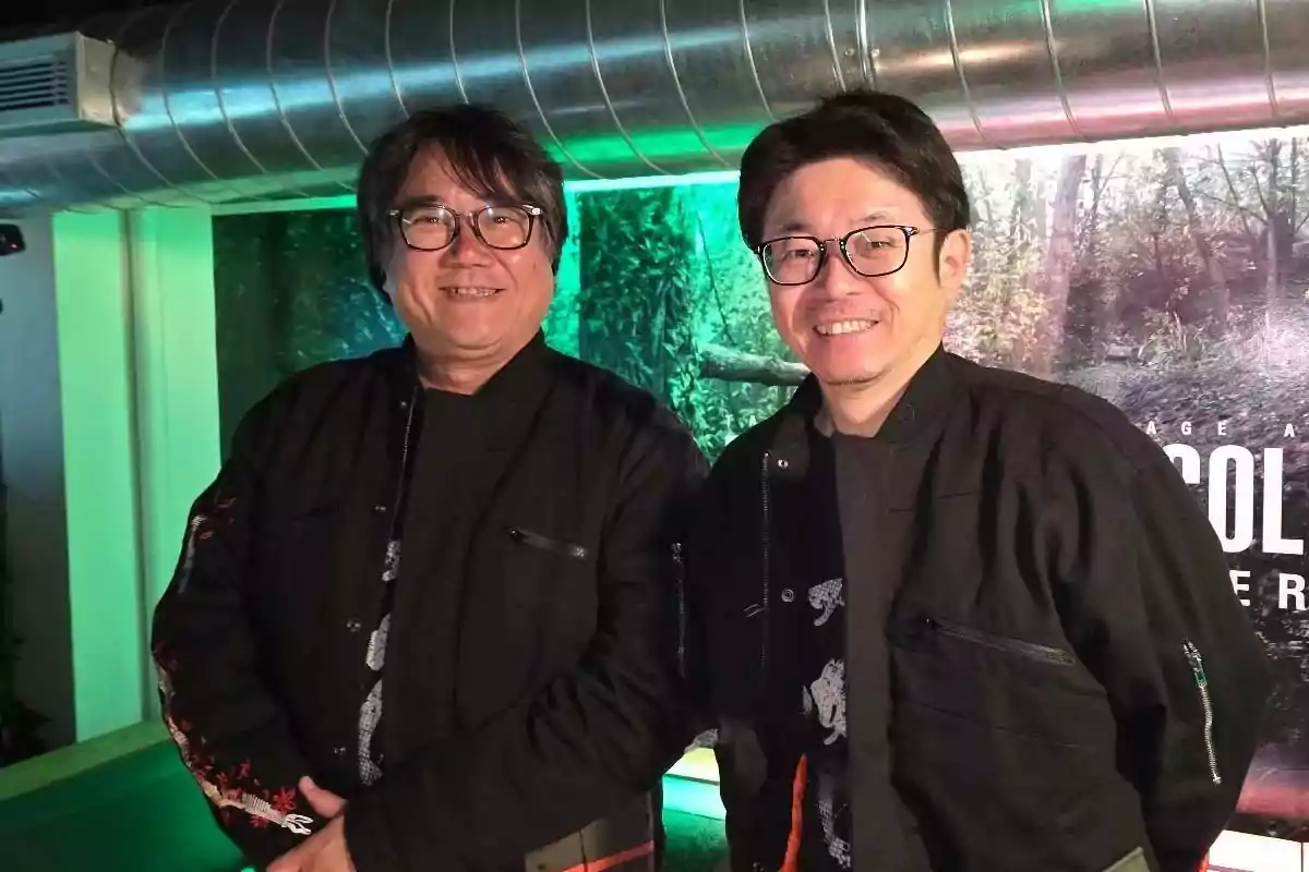 Two smiling men wearing glasses and black jackets pose together indoors with green lights and a nature background. Two smiling men wearing glasses and black jackets pose together indoors with green lights and a nature background.
