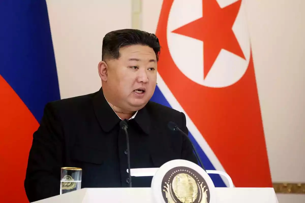 Short dark-haired man speaking at a podium with microphones, with North Korean and Russian flags visible in the background.