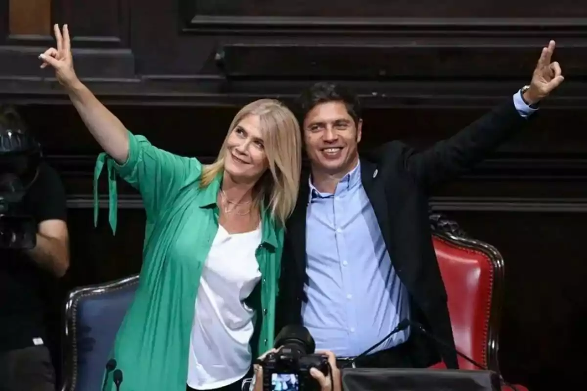 Two people smile and raise their arms making the victory sign while posing for a photo in a formal setting. Two people smile and raise their arms making the victory sign while posing for a photo in a formal setting.