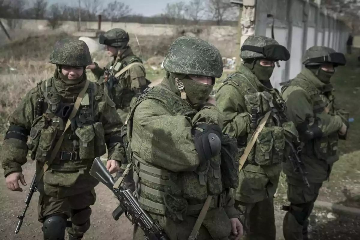 Soldiers in green camouflage uniforms and military gear walking in formation along a dirt road next to a fence.