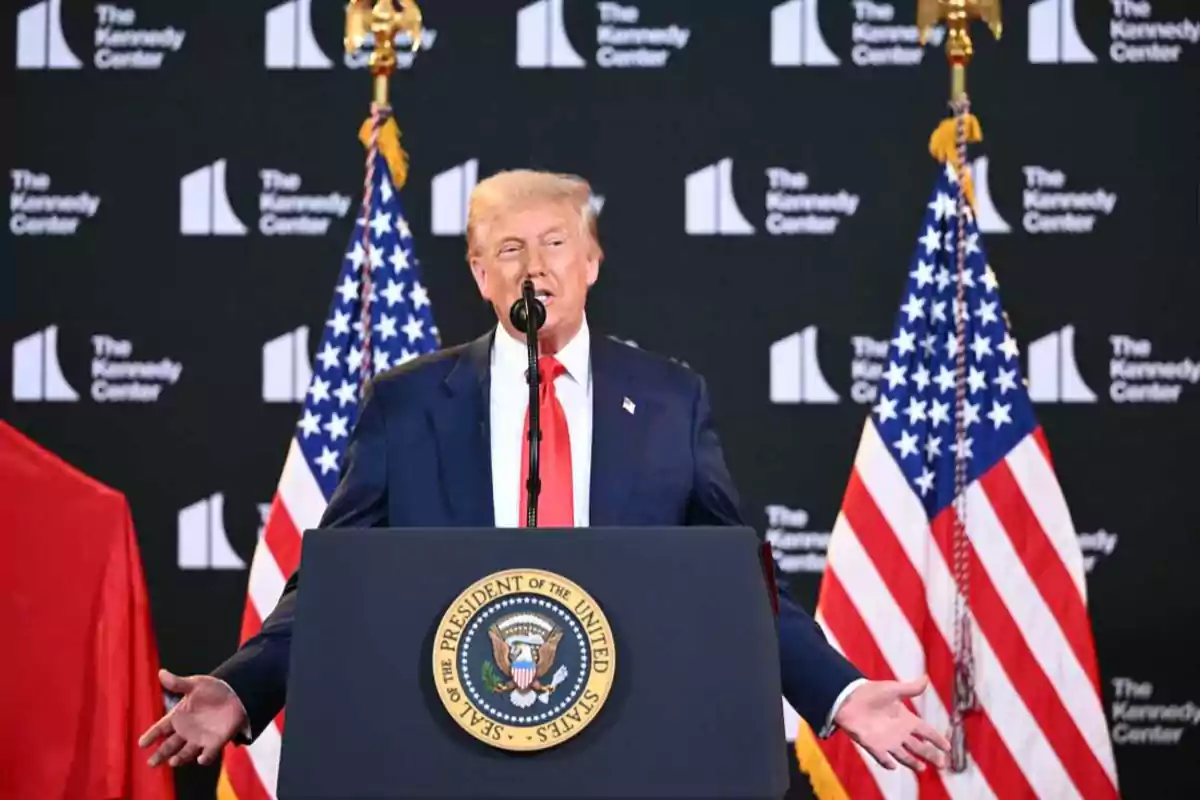 Hombre de traje azul y corbata roja hablando en un podio presidencial con dos banderas de Estados Unidos al fondo y el logo de The Kennedy Center en la pared. Hombre de traje azul y corbata roja hablando en un podio presidencial con dos banderas de Estados Unidos al fondo y el logo de The Kennedy Center en la pared.