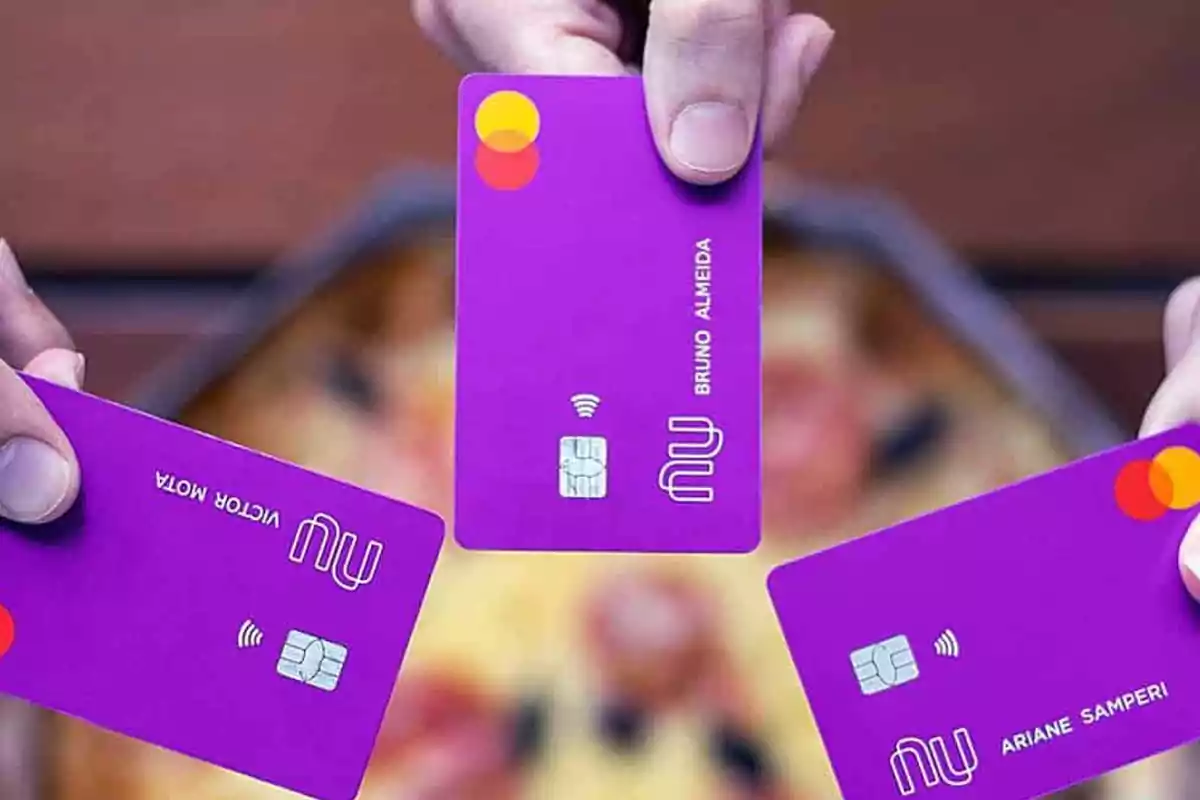 Tres personas sostienen tarjetas bancarias moradas de la marca Nubank sobre una mesa con pizza desenfocada al fondo Tres personas sostienen tarjetas bancarias moradas de la marca Nubank sobre una mesa con pizza desenfocada al fondo