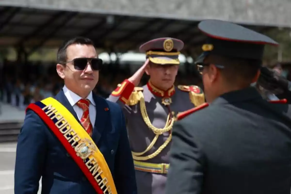 Un hombre con banda presidencial y gafas oscuras está acompañado por oficiales militares en un evento formal al aire libre Un hombre con banda presidencial y gafas oscuras está acompañado por oficiales militares en un evento formal al aire libre