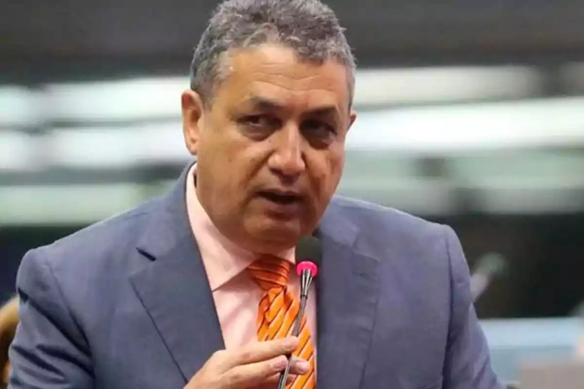 Hombre de cabello canoso con traje azul y corbata naranja hablando frente a un micrófono