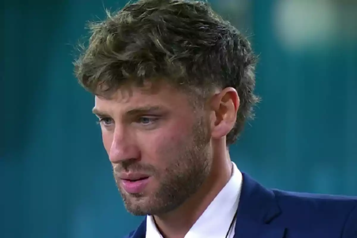 Hombre joven con barba y cabello rizado luce pensativo y viste un traje azul con camisa blanca sobre un fondo desenfocado azul Hombre joven con barba y cabello rizado luce pensativo y viste un traje azul con camisa blanca sobre un fondo desenfocado azul