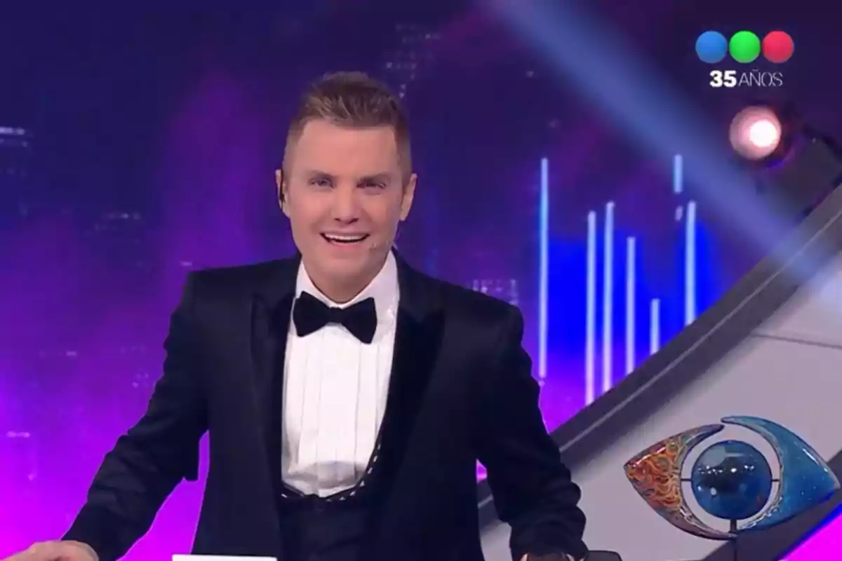Man in a tuxedo hosting a TV show with a purple background and a channel logo in the top right corner