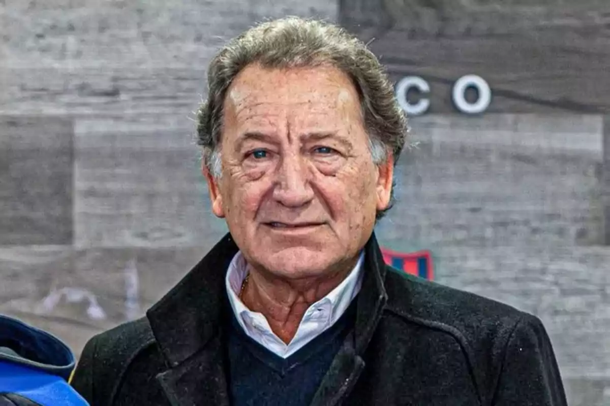 Older man with gray hair and a serious expression, wearing a dark coat and white jersey in front of a gray background Older man with gray hair and a serious expression, wearing a dark coat and white jersey in front of a gray background