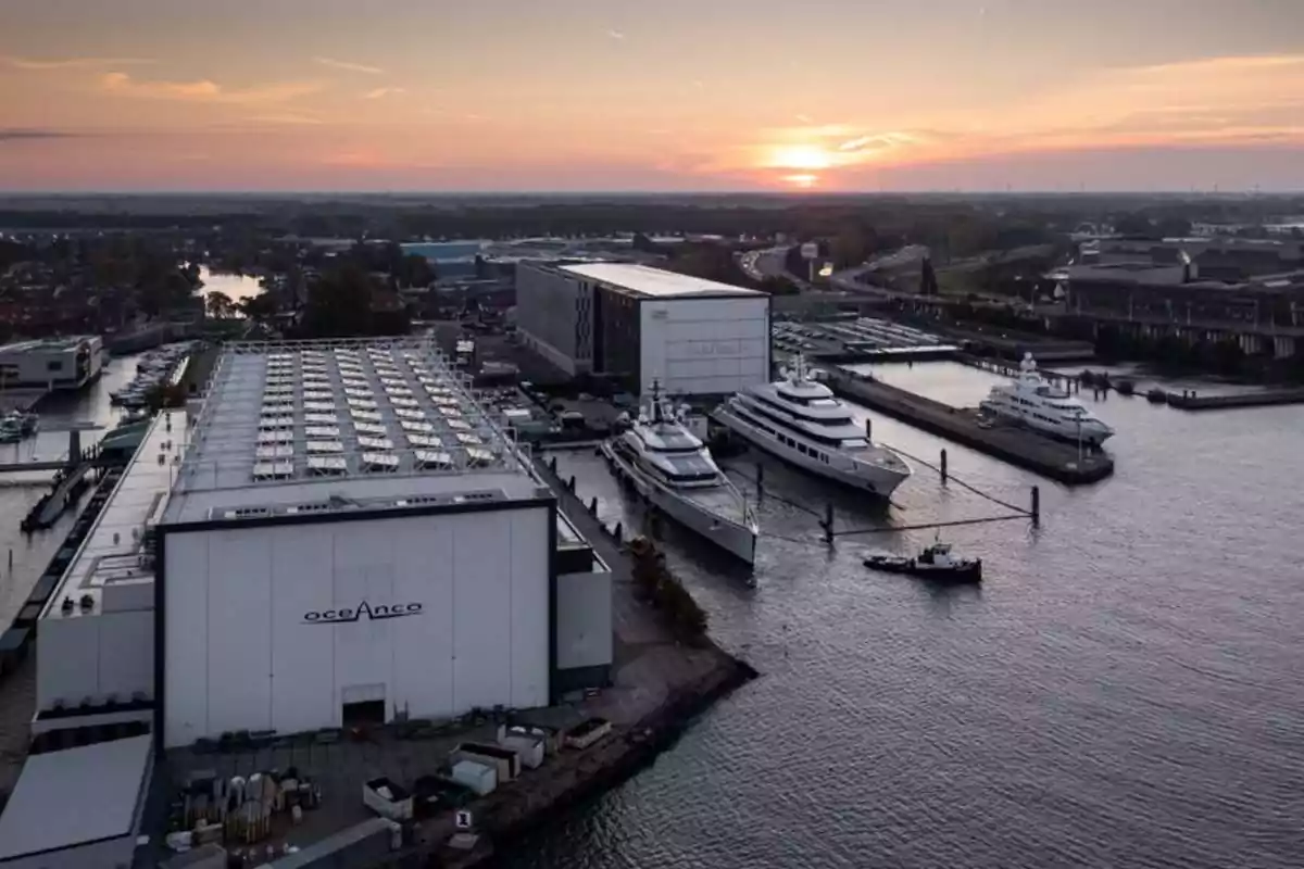 Astillero con varios yates de lujo atracados junto a grandes naves industriales al atardecer