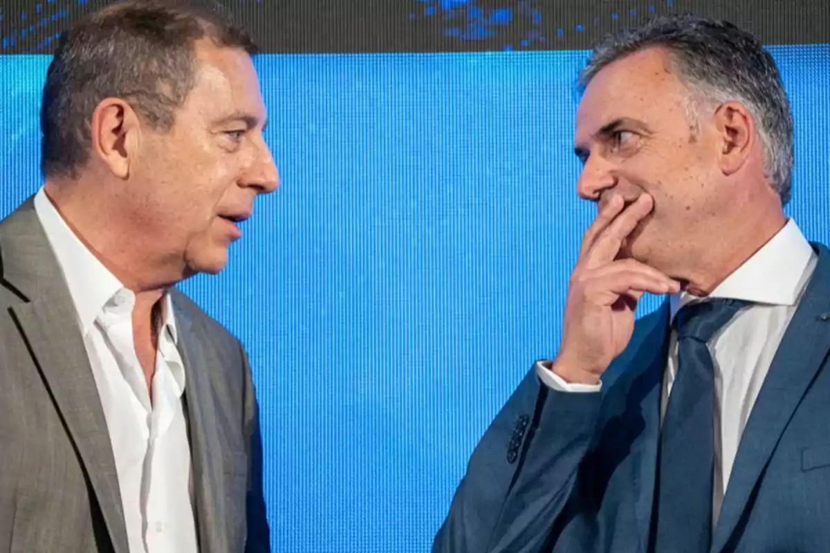 Two men in suits talk in front of a blue background Two men in suits talk in front of a blue background