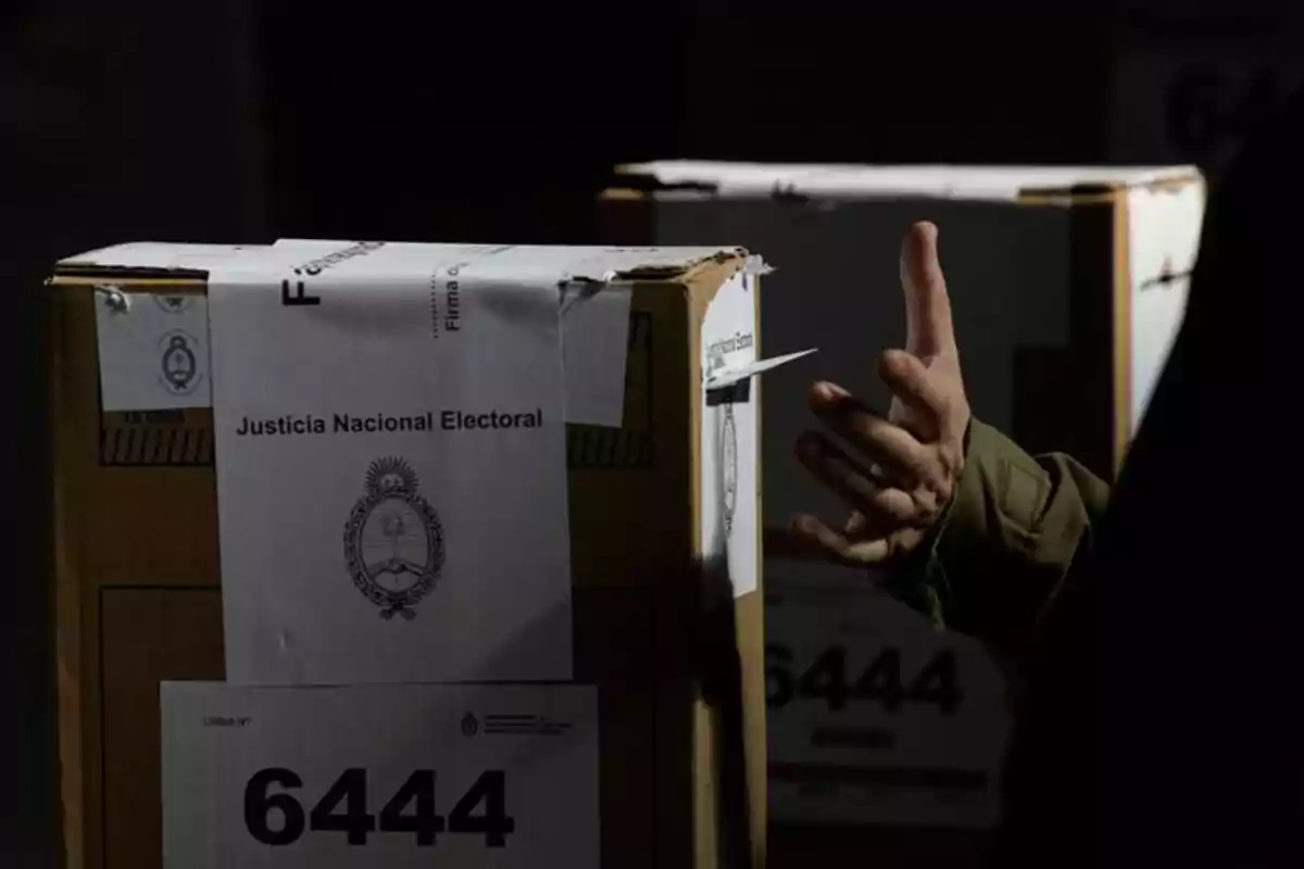 Una mano junto a una urna electoral de cartón con el sello de la Justicia Nacional Electoral de Argentina y el número 6444 en primer plano