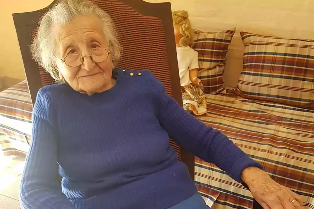 Older woman with glasses and a blue sweater sitting on a chair next to a bed with checkered sheets and a blonde doll in the background