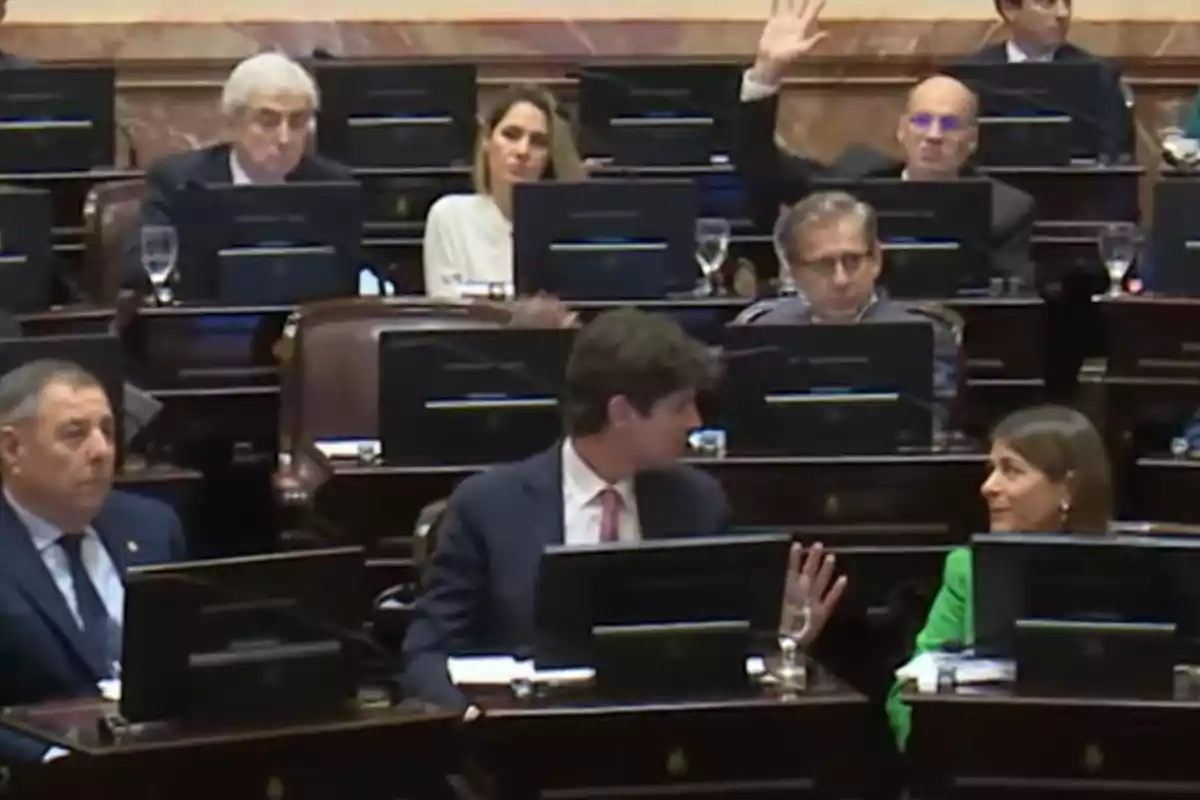 Personas sentadas en un recinto legislativo con computadoras frente a ellas, una persona al fondo levanta la mano Personas sentadas en un recinto legislativo con computadoras frente a ellas, una persona al fondo levanta la mano