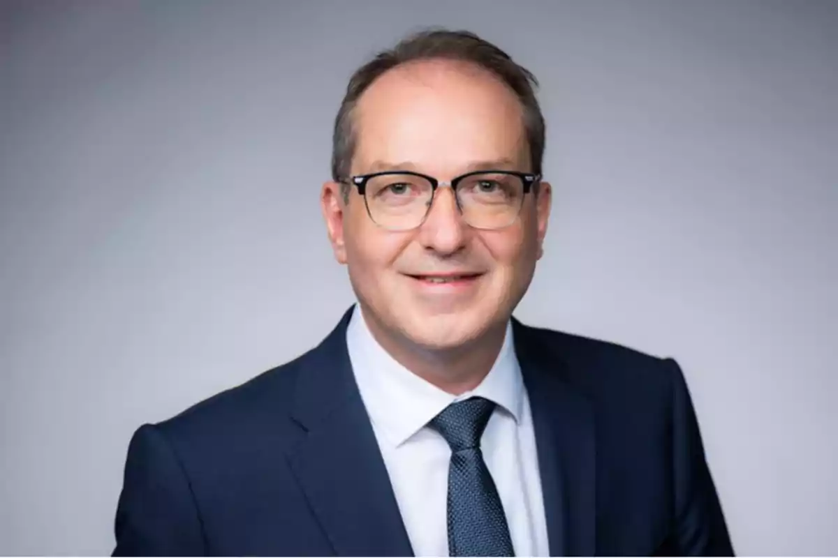 Middle-aged man with glasses and a blue suit smiling in front of a light gray background Middle-aged man with glasses and a blue suit smiling in front of a light gray background