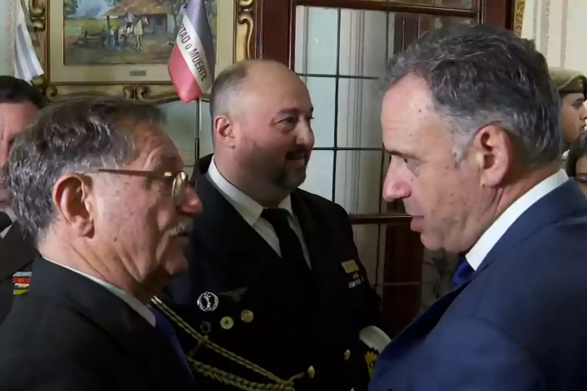 Tres hombres conversan en un ambiente formal con uniformes y trajes, al fondo se observa una bandera y un cuadro enmarcado.