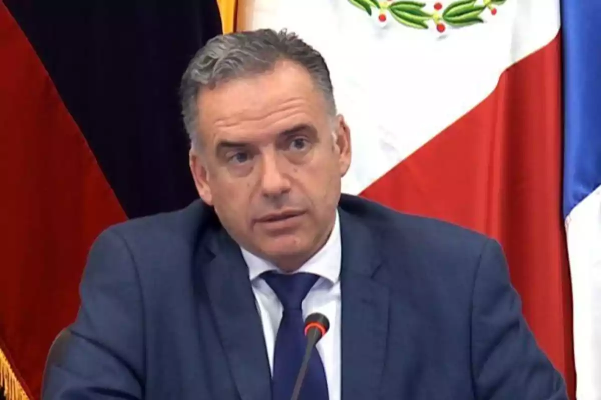 Man in a blue suit speaking in front of a microphone with flags in the background Man in a blue suit speaking in front of a microphone with flags in the background