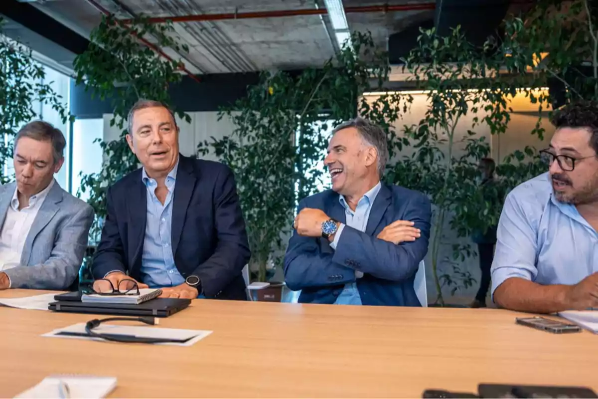 Four men sitting at a conference table talk and smile in a modern office with plants in the background. Four men sitting at a conference table talk and smile in a modern office with plants in the background.