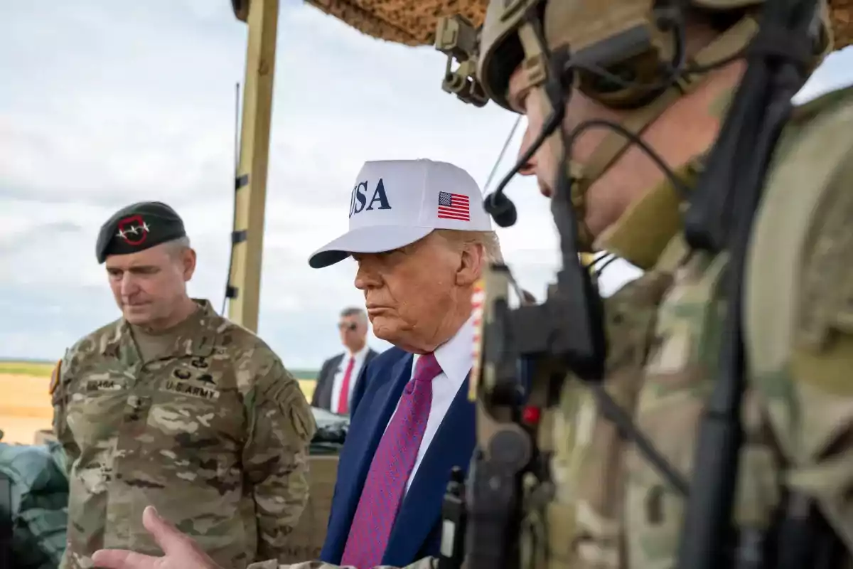 A man wearing a white cap and a blue suit is accompanied by two soldiers in camouflage uniforms in an outdoor setting.
