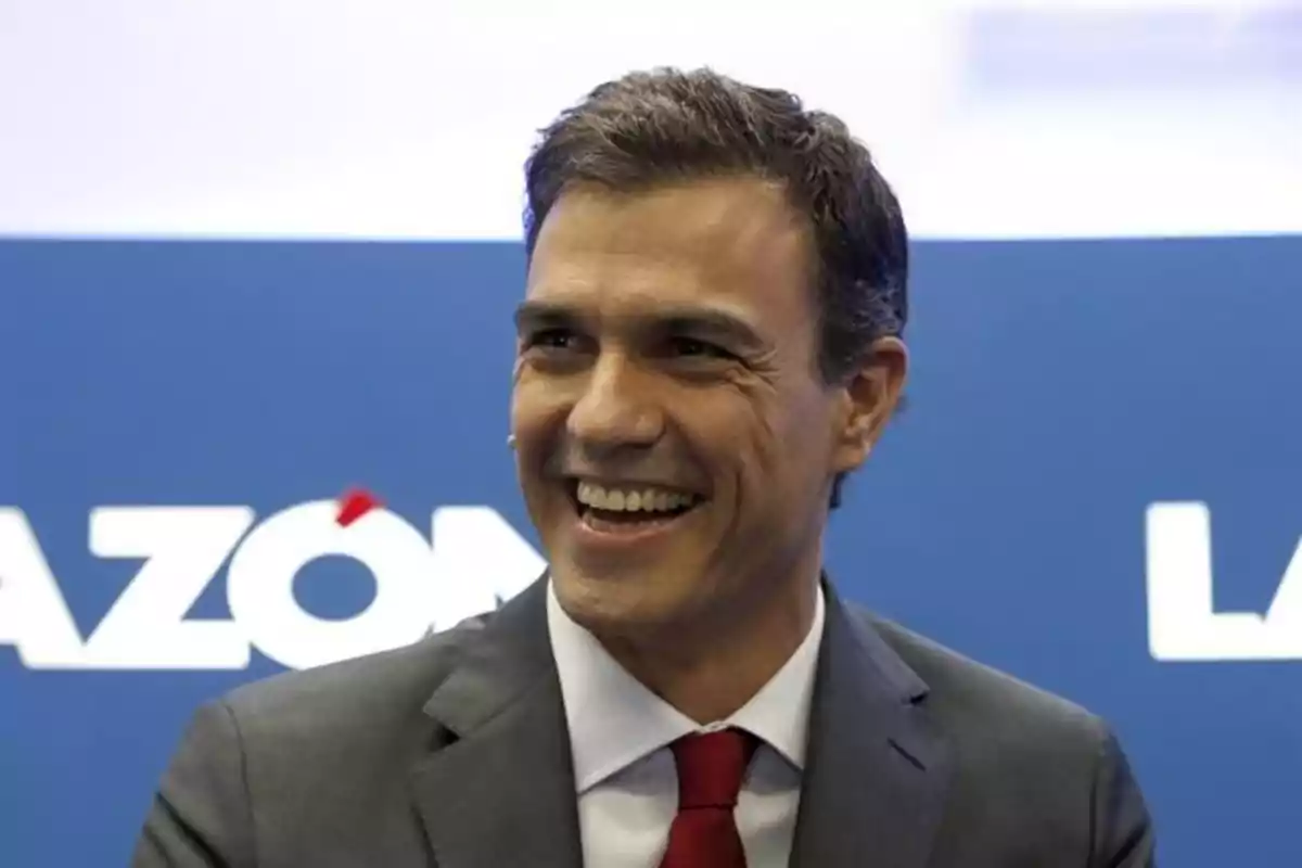 Short dark-haired man in a gray suit and red tie smiling in front of a blue background with white letters