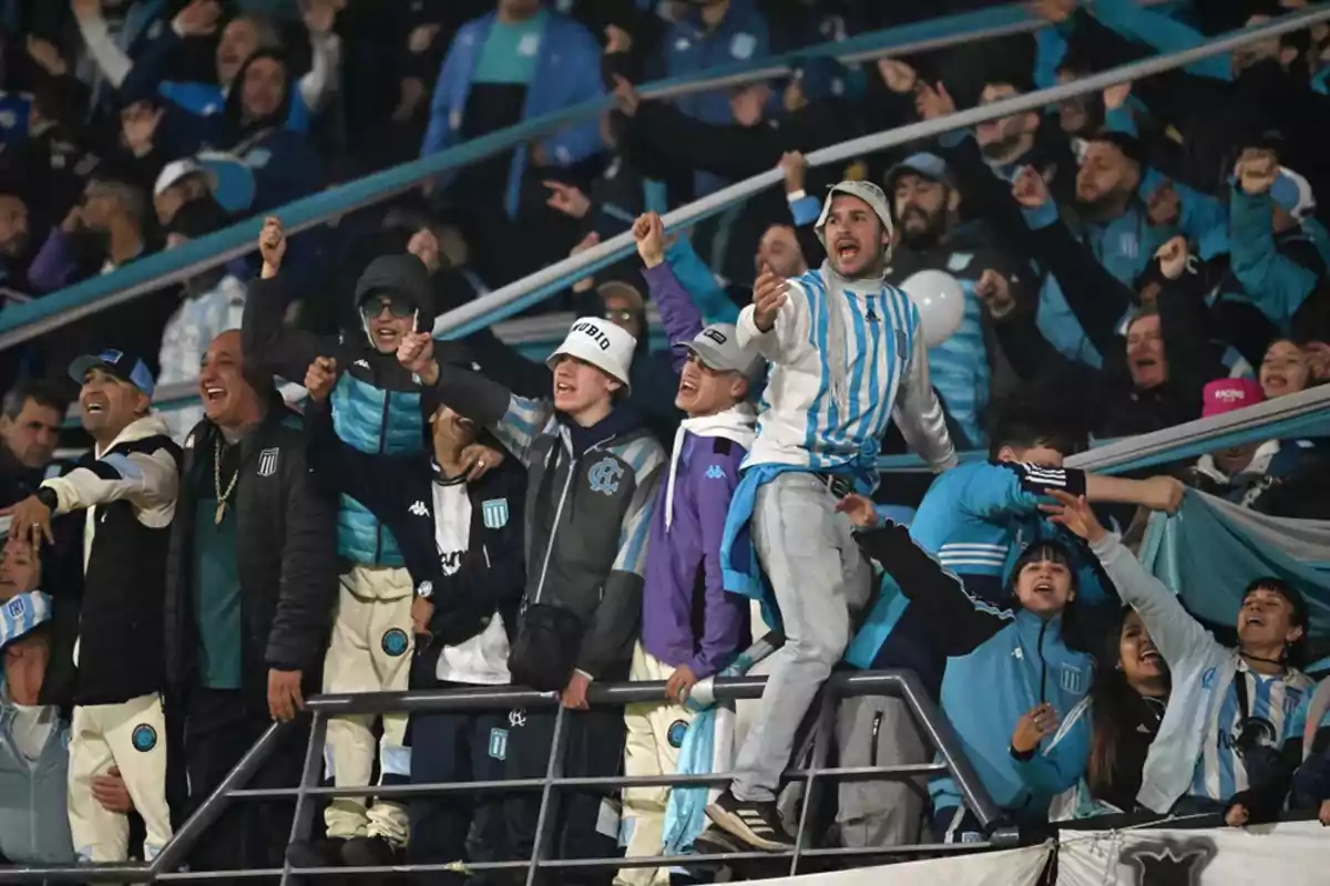 Racing Club fans celebrate and sing in the stands of a stadium, dressed in light blue and white clothing.