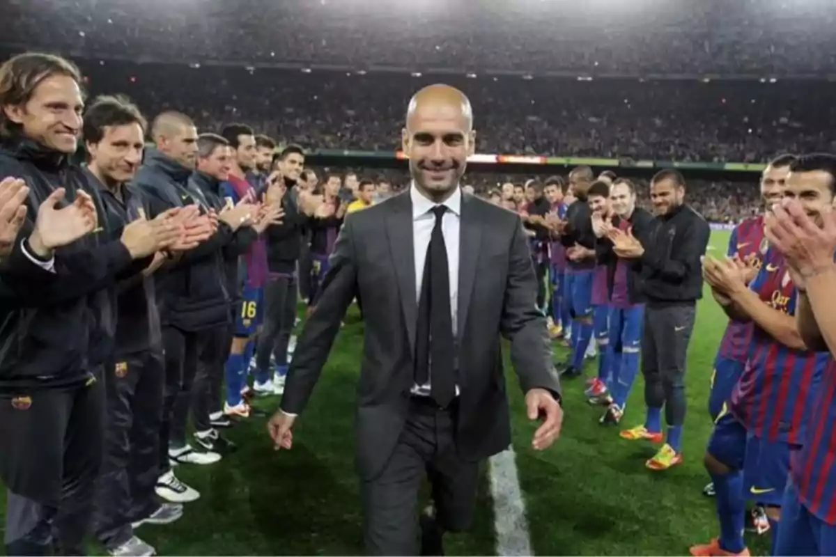 Un hombre con traje camina sonriente por una fila de jugadores de fútbol que lo aplauden en un estadio lleno de público Un hombre con traje camina sonriente por una fila de jugadores de fútbol que lo aplauden en un estadio lleno de público