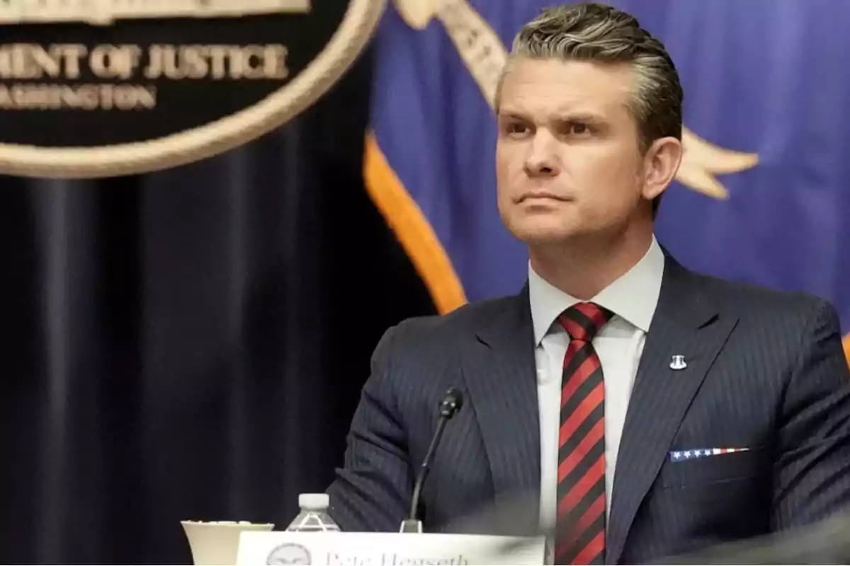 Man in a dark suit with a red and black tie sitting at a table during an official event with a background of flags and a partial Department of Justice sign Man in a dark suit with a red and black tie sitting at a table during an official event with a background of flags and a partial Department of Justice sign