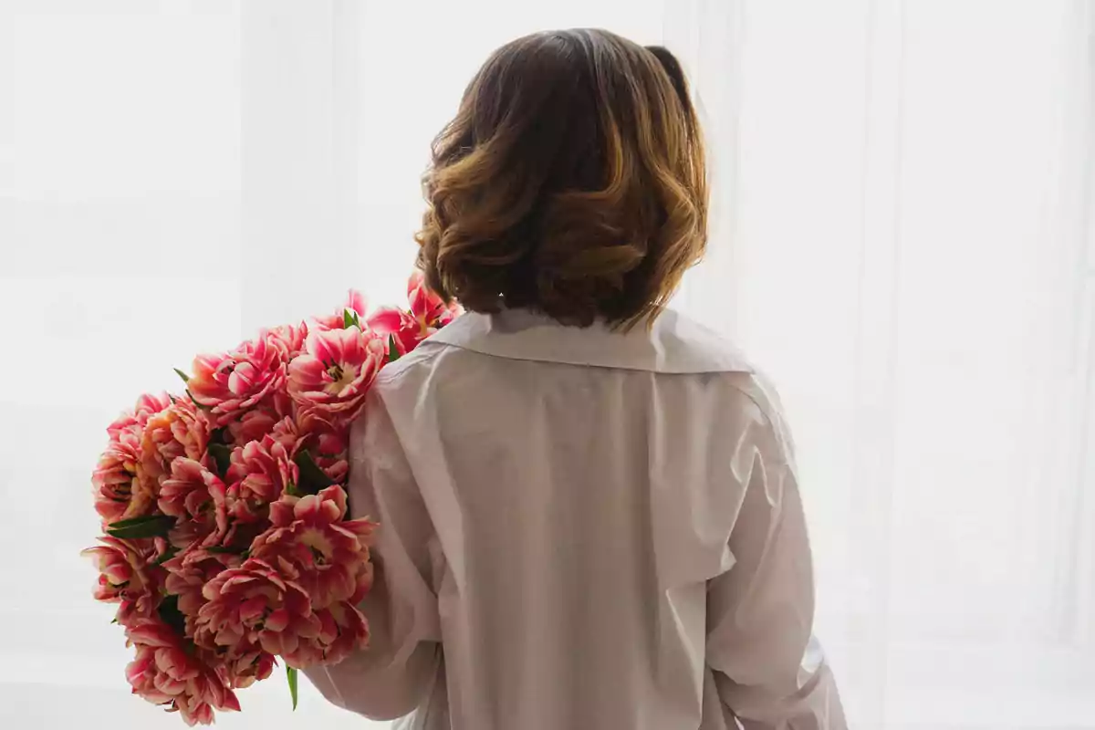 Persona de espaldas con camisa blanca sosteniendo un gran ramo de flores rosas frente a una ventana iluminada