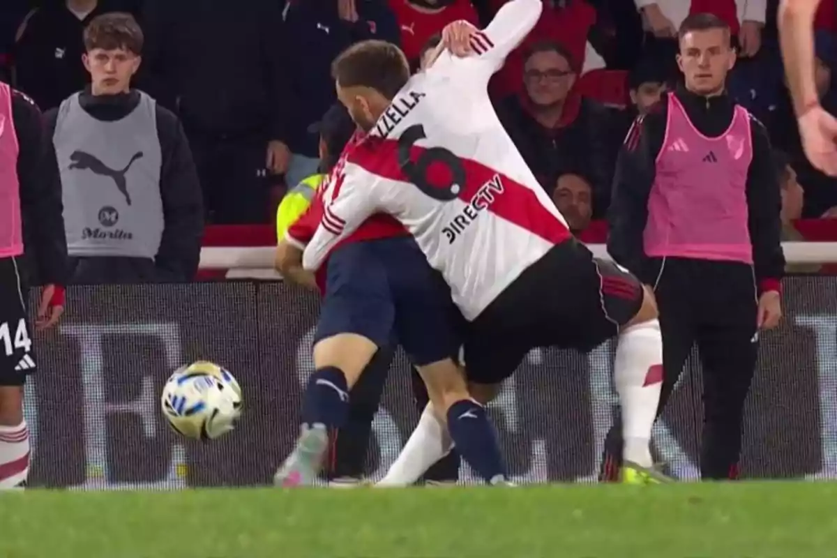 Jugador de River Plate comete una falta sujetando a un rival durante un partido de fútbol mientras otros jugadores observan desde la banda