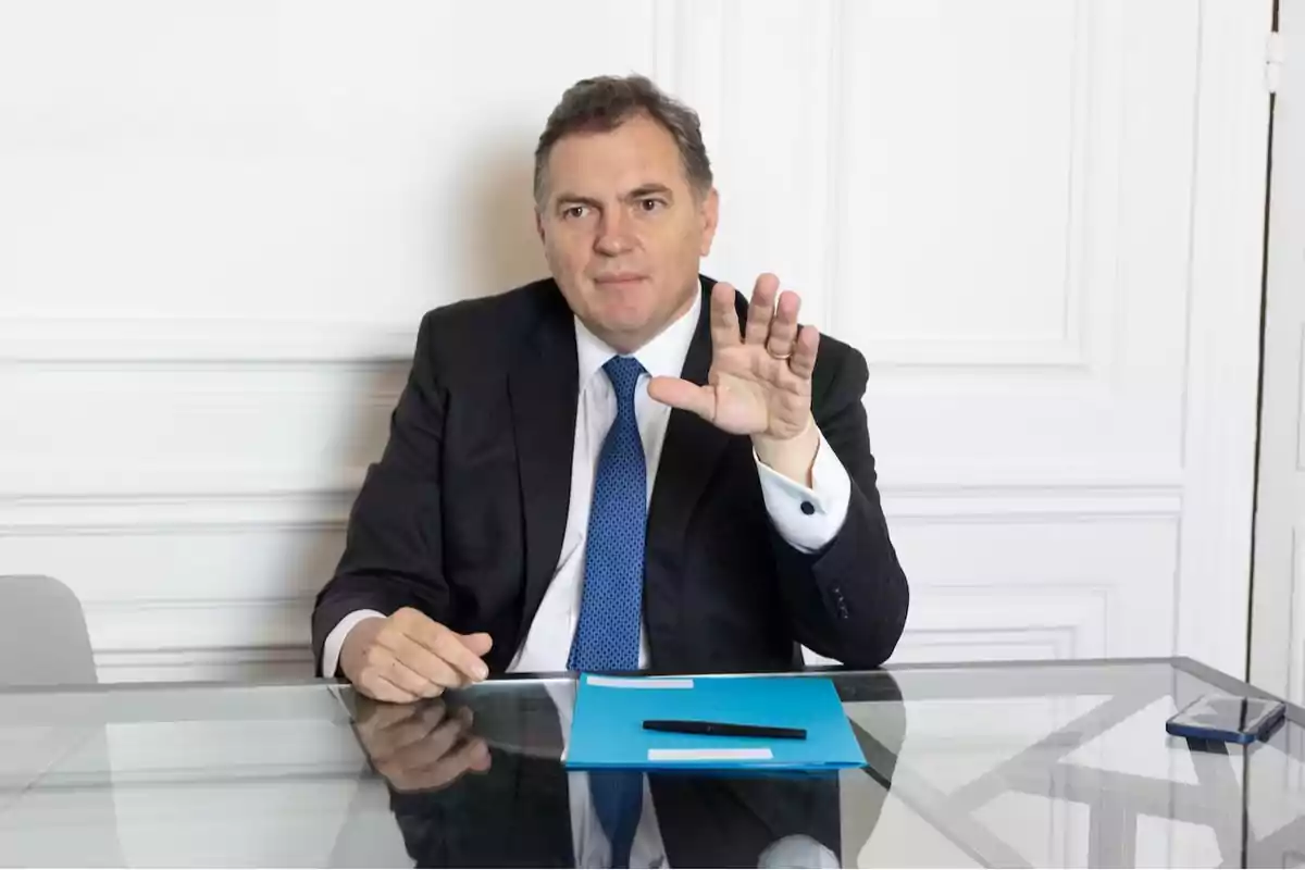 Hombre de traje sentado en una mesa de cristal con una carpeta azul y un bolígrafo, levantando la mano mientras mira al frente Hombre de traje sentado en una mesa de cristal con una carpeta azul y un bolígrafo, levantando la mano mientras mira al frente