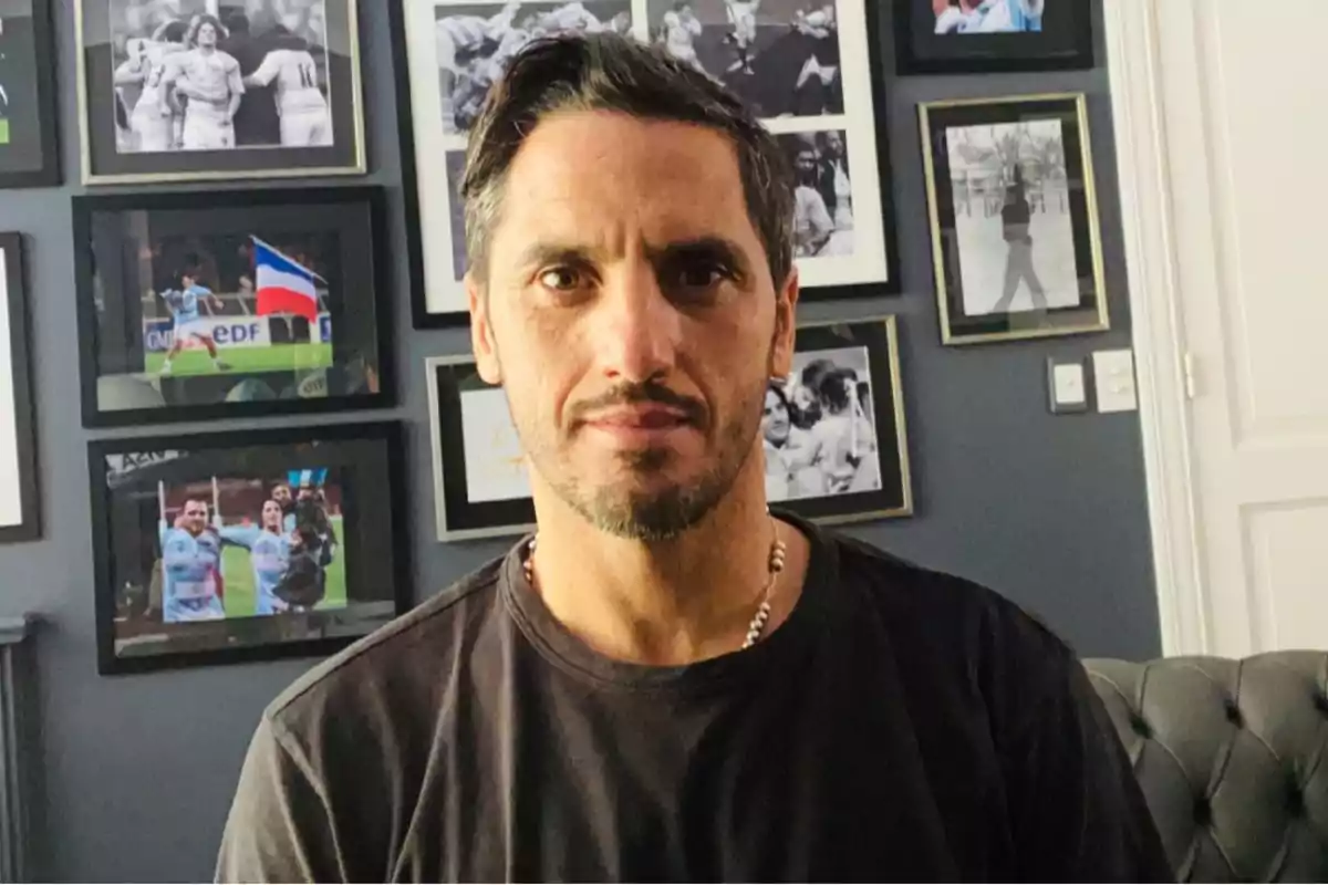Hombre de cabello corto y barba ligera sentado en una habitación con varias fotos enmarcadas en la pared detrás de él