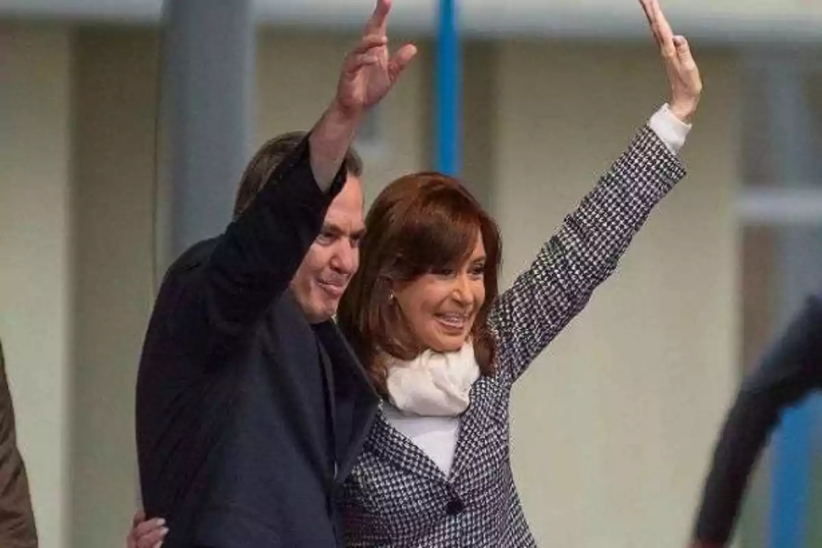 Two people smile and wave with their arms raised while hugging.