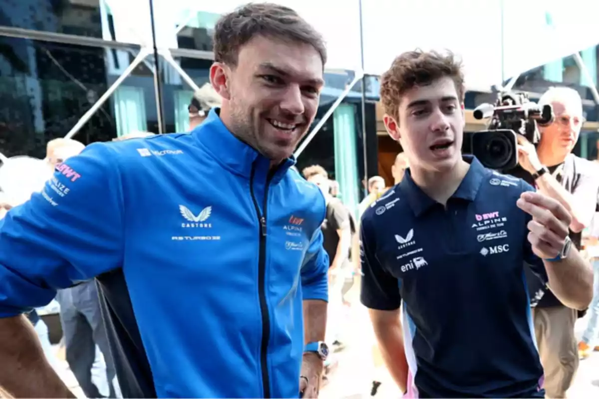 Two men in blue sportswear talk and smile while a camera records them in an outdoor setting with more people in the background. Two men in blue sportswear talk and smile while a camera records them in an outdoor setting with more people in the background.