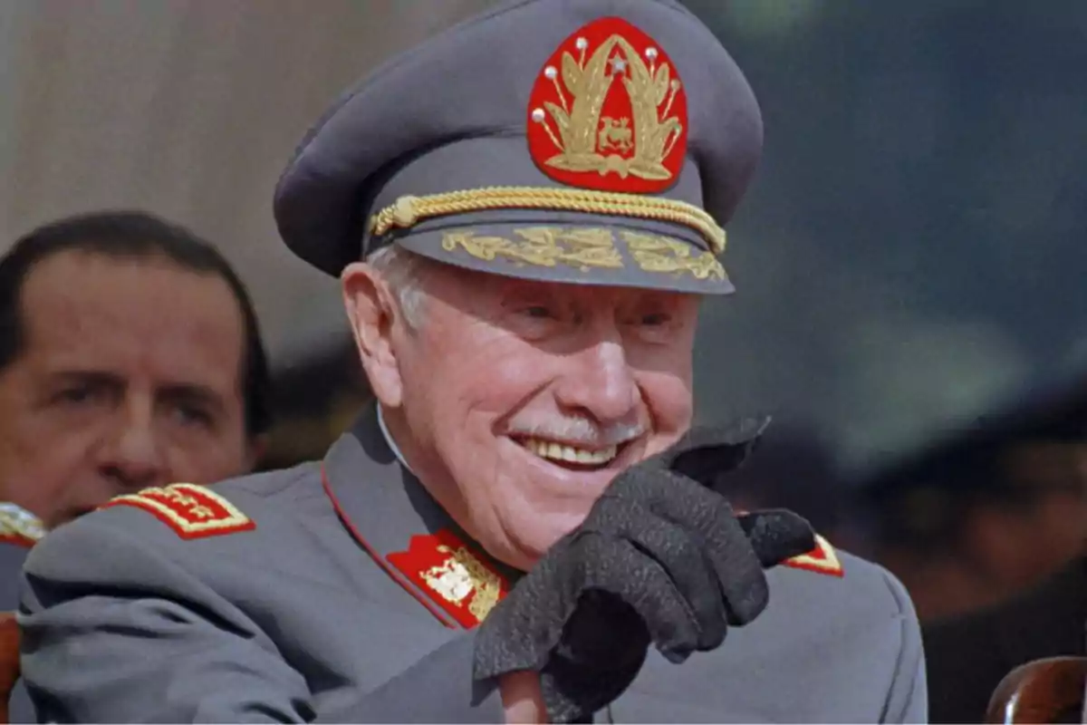 Elderly man in a gray military uniform and decorated cap smiling and pointing with a black glove