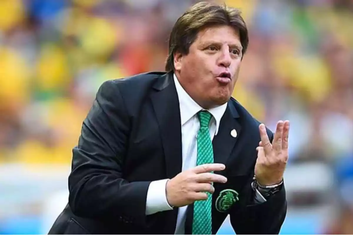 Entrenador de fútbol con traje oscuro y corbata verde haciendo una seña con la mano durante un partido