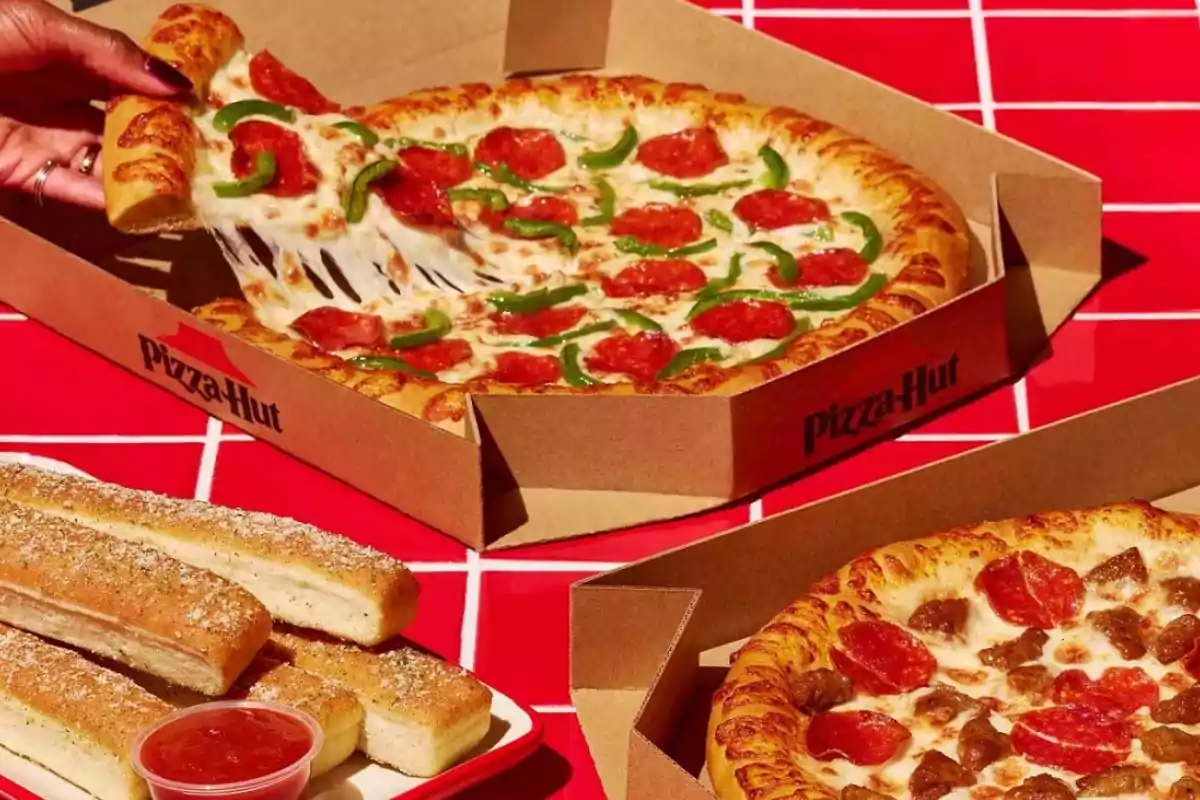 Una mano toma una rebanada de pizza con queso derretido, pepperoni y pimientos verdes en una caja de Pizza Hut, junto a otra pizza y palitos de pan con salsa marinara sobre una mesa roja.