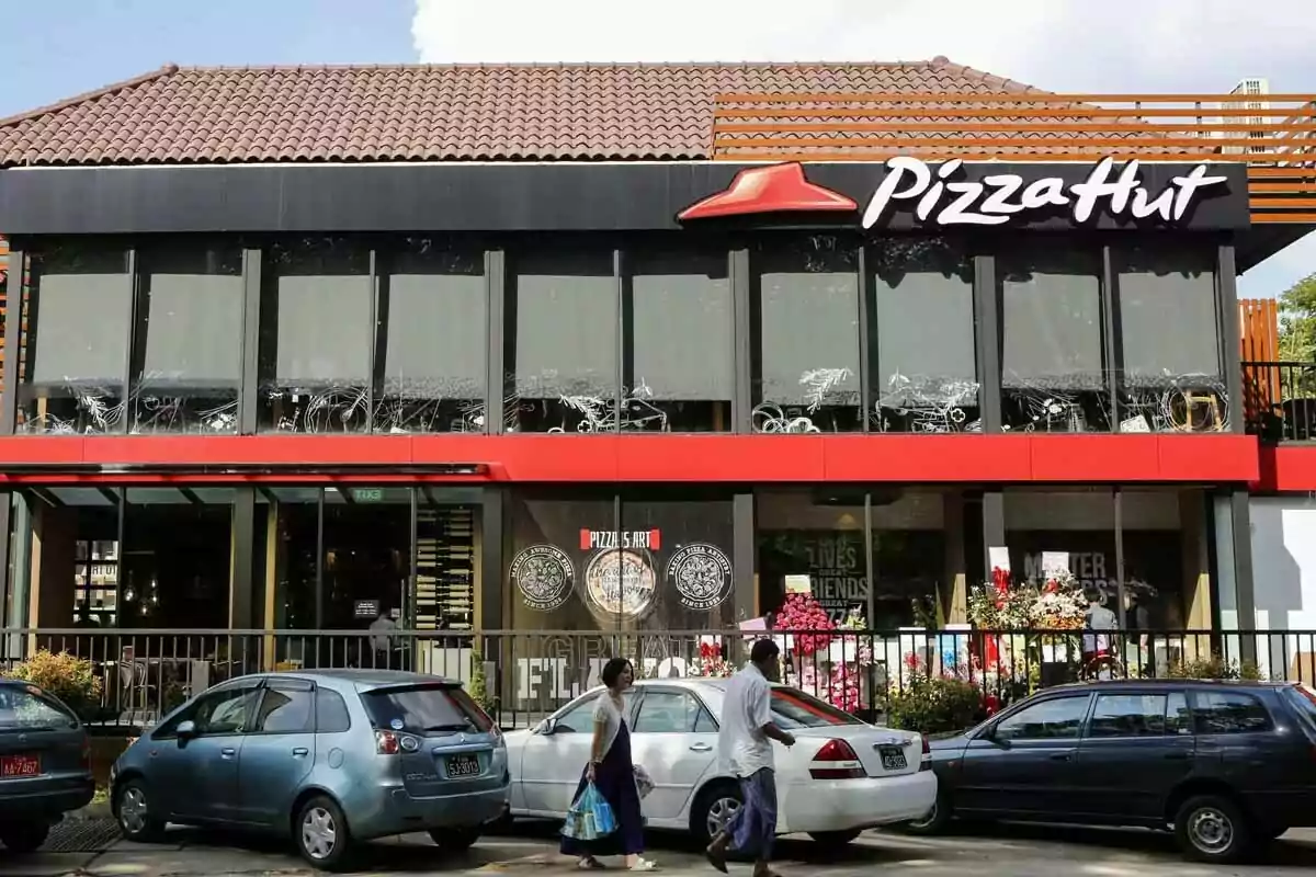 Fachada de un restaurante Pizza Hut con varios autos estacionados al frente y dos personas caminando por la acera