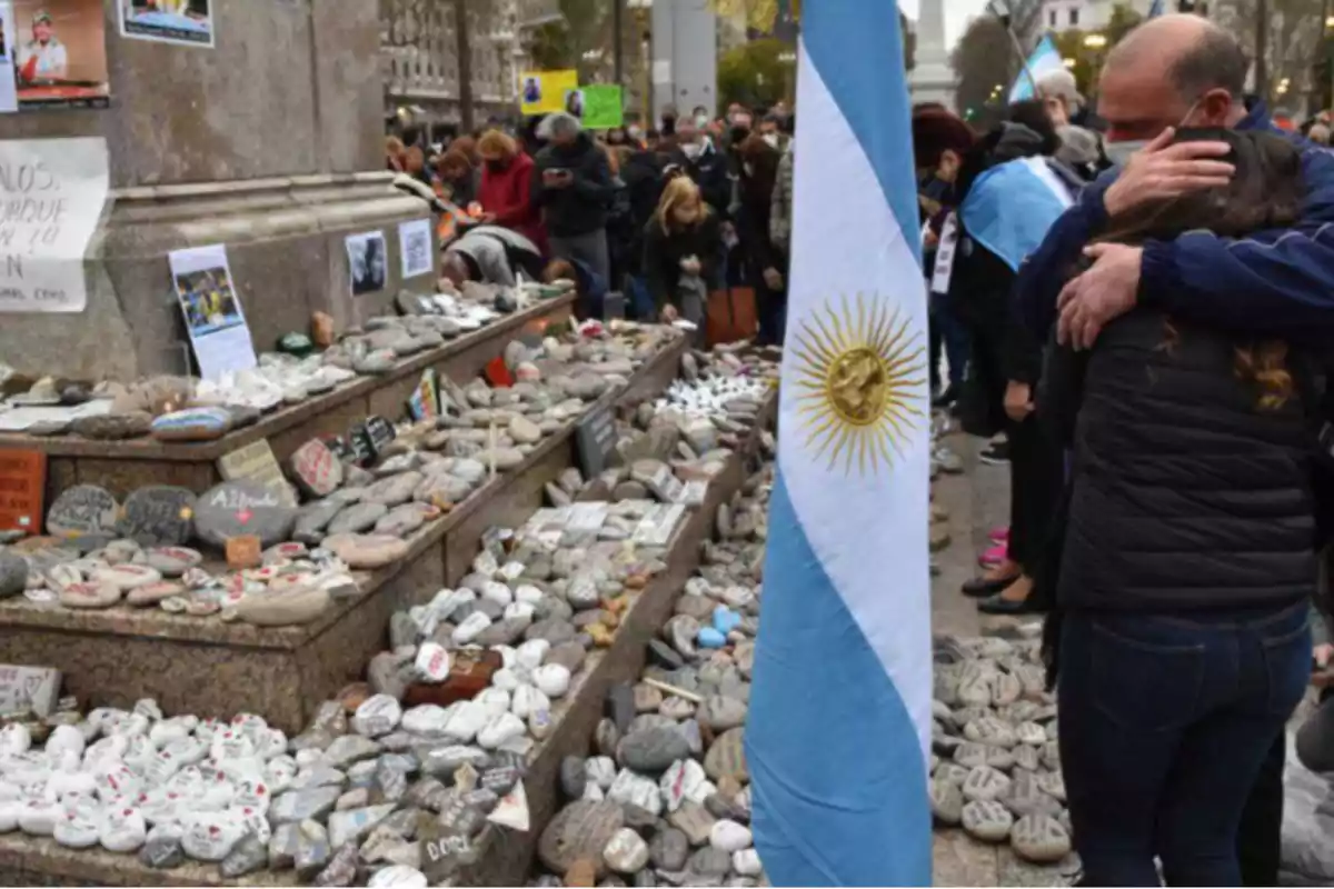 Personas reunidas en un monumento cubierto de piedras con mensajes y una bandera argentina en primer plano