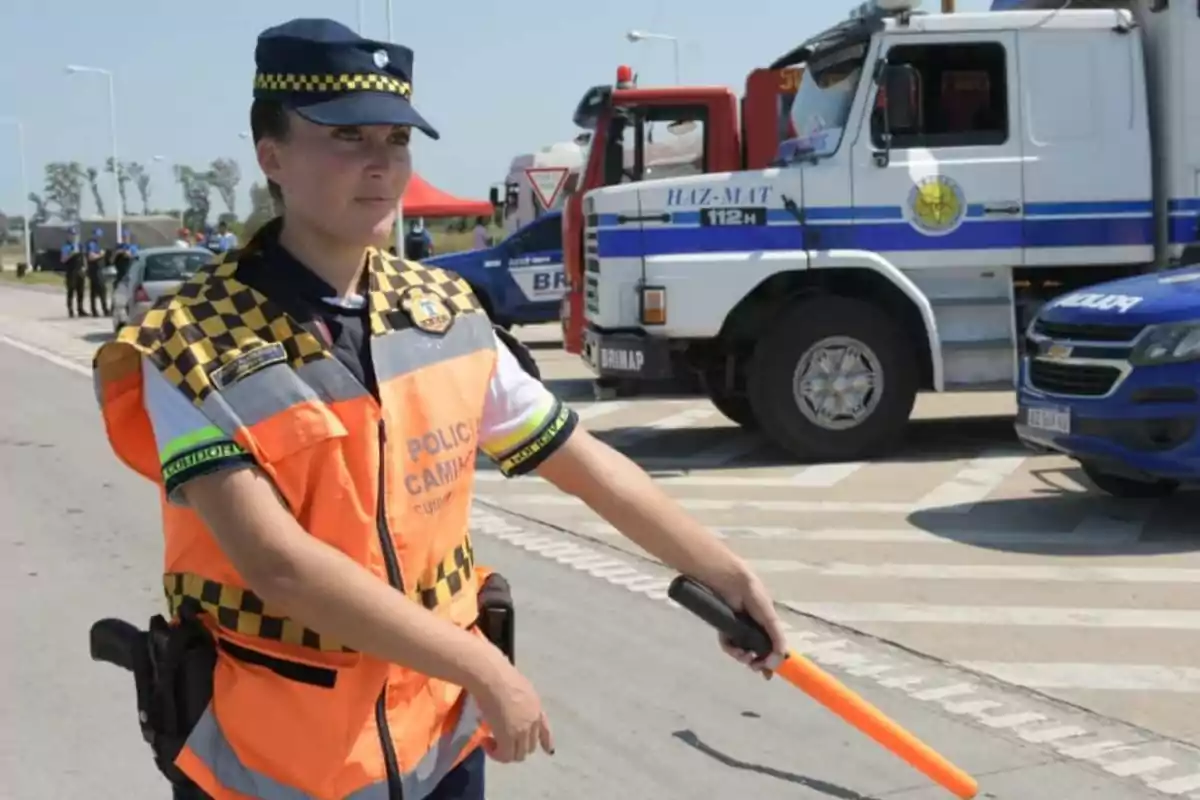Agente de policía de tránsito con chaleco naranja dirigiendo el tráfico en una carretera junto a vehículos oficiales y camiones de emergencia